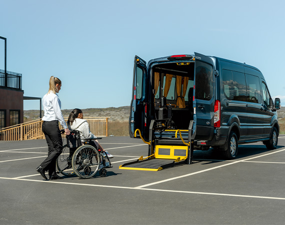 A female driver is pushing young woman in a wheelchair towards a parked vehicle with a wheelchair lift folded out from the back.