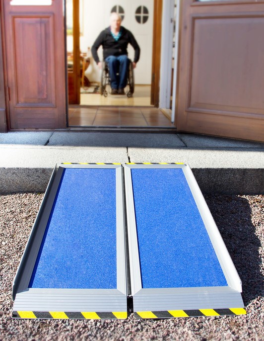 A wheelchair ramp leading up a step into a house, a man in the back moving towards the door. 