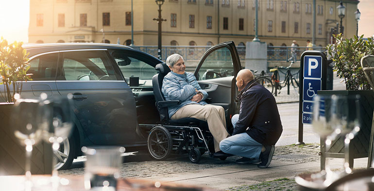 Elderly man assisting elderly woman in exiting a car using a transfer wheelchair.