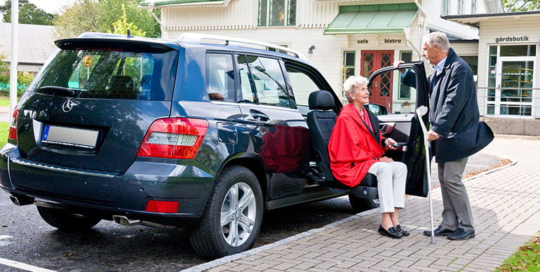 Elderly man assisting an elderly woman exiting a car using a seat lift. 
