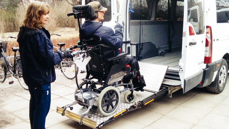 Eine Frau hilft einem Mann im Rollstuhl mit einem Lift in einen Van