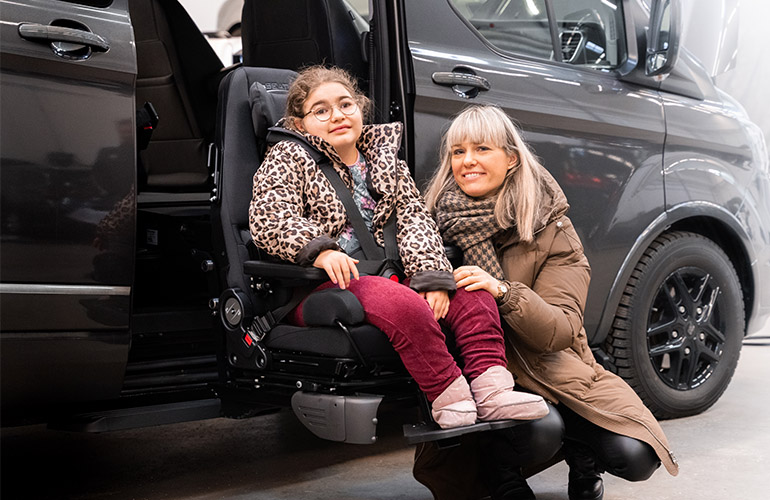 Girl sitting in a specially adapted seat attached to the Turny Evo seat lift outside of a car and her mother crouching next to her