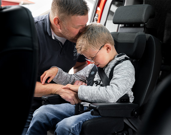 A young boy seated in a car seat with his father leaning in from the door opening holding the boy's hand and kissing his head.