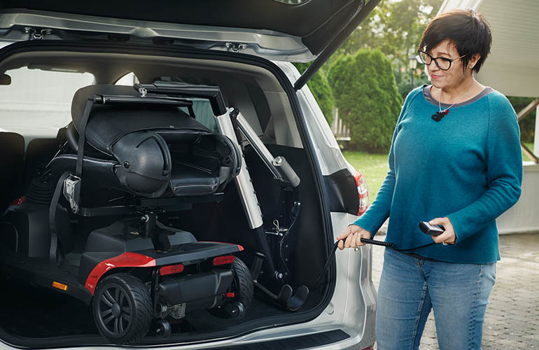 Der Kofferraum eines Autos mit einem Elektromobil, das an einem Carolift 100 befestigt ist, und eine Frau, die neben dem Auto steht und eine Fernbedienung hält.